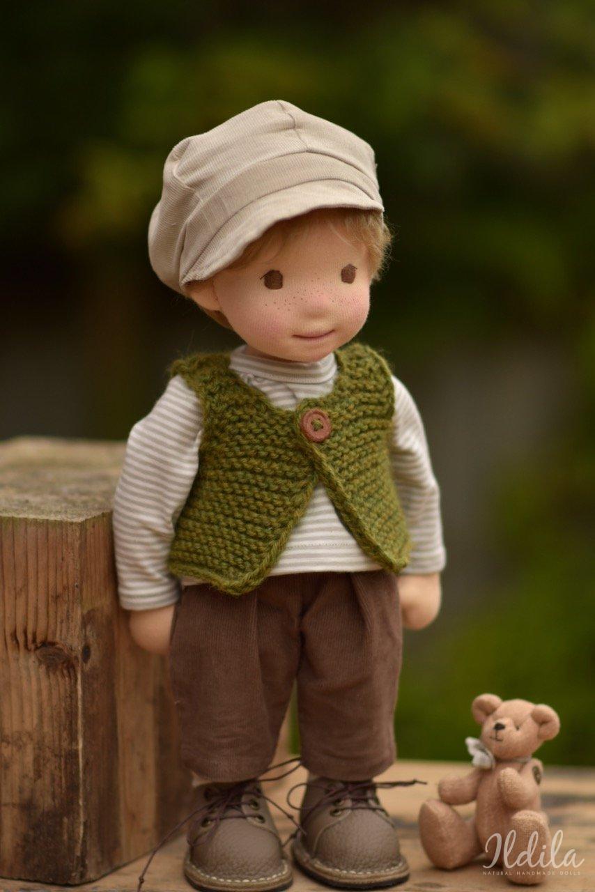 Handgemaakte pop met beige muts, gestreept shirt, groen vest, bruine broek en schoenen naast teddybeer.