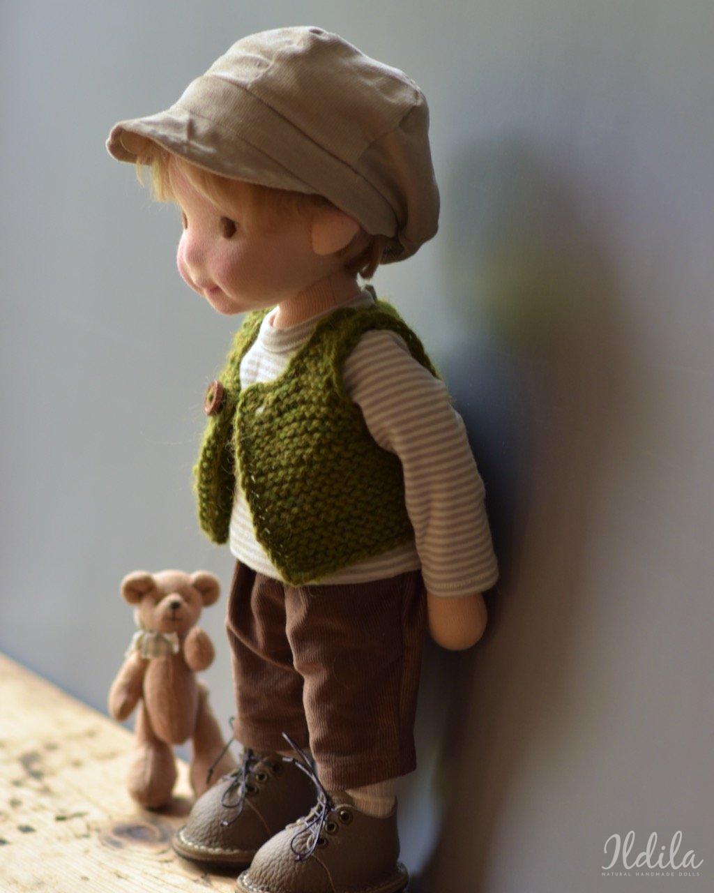 Handgemaakte pop in beige hoed, gestreept shirt, groen vest, bruine broek en schoenen met teddybeer in de buurt.