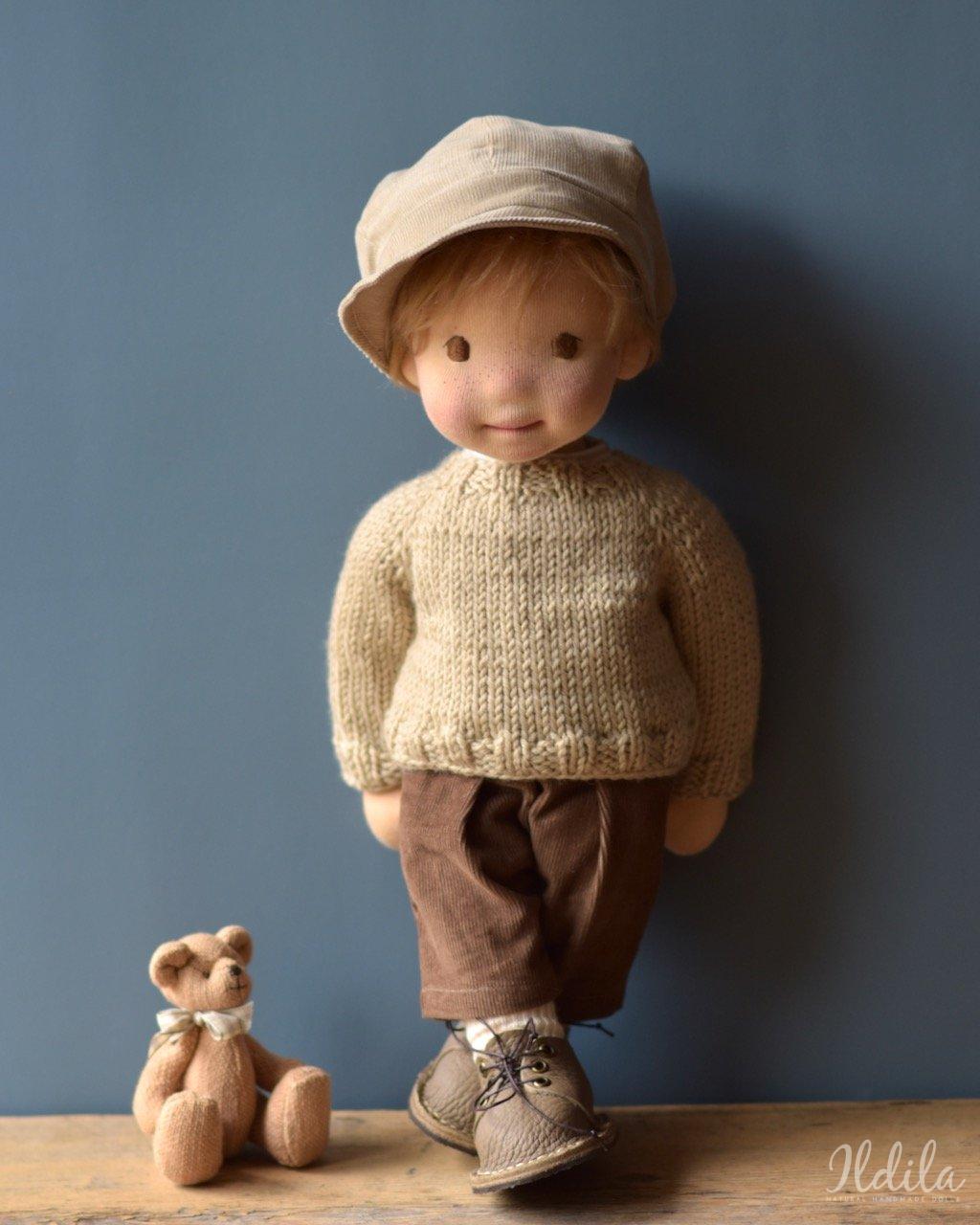 Handgemaakte pop in beige trui, bruine broek, hoed en schoenen met kleine teddybeer op houten ondergrond.